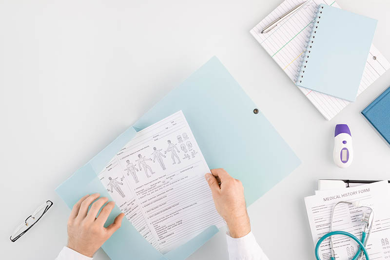 Hands of doctor putting medical document into folder with other files while sitting by workplace with tools, notebooks and papers